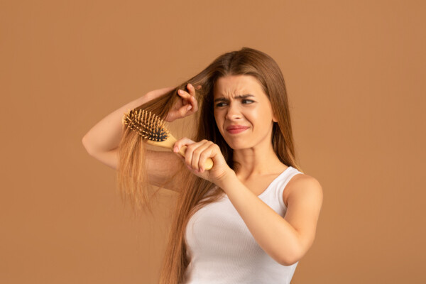 Annoyed young woman having problem brushing her long hair on brown studio background